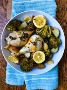A bowl with chicken drumsticks, broccoli florets, baby red potatoes, lemon slices, and garlic cloves for Sheet Pan Lemon Garlic Chicken. Cooking With Fudge