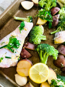 A closeup view of a sheet pan with chicken drumsticks, broccoli florets, baby red potatoes, lemon slices, and garlic cloves for Sheet Pan Lemon Garlic Chicken. Cooking With Fudge