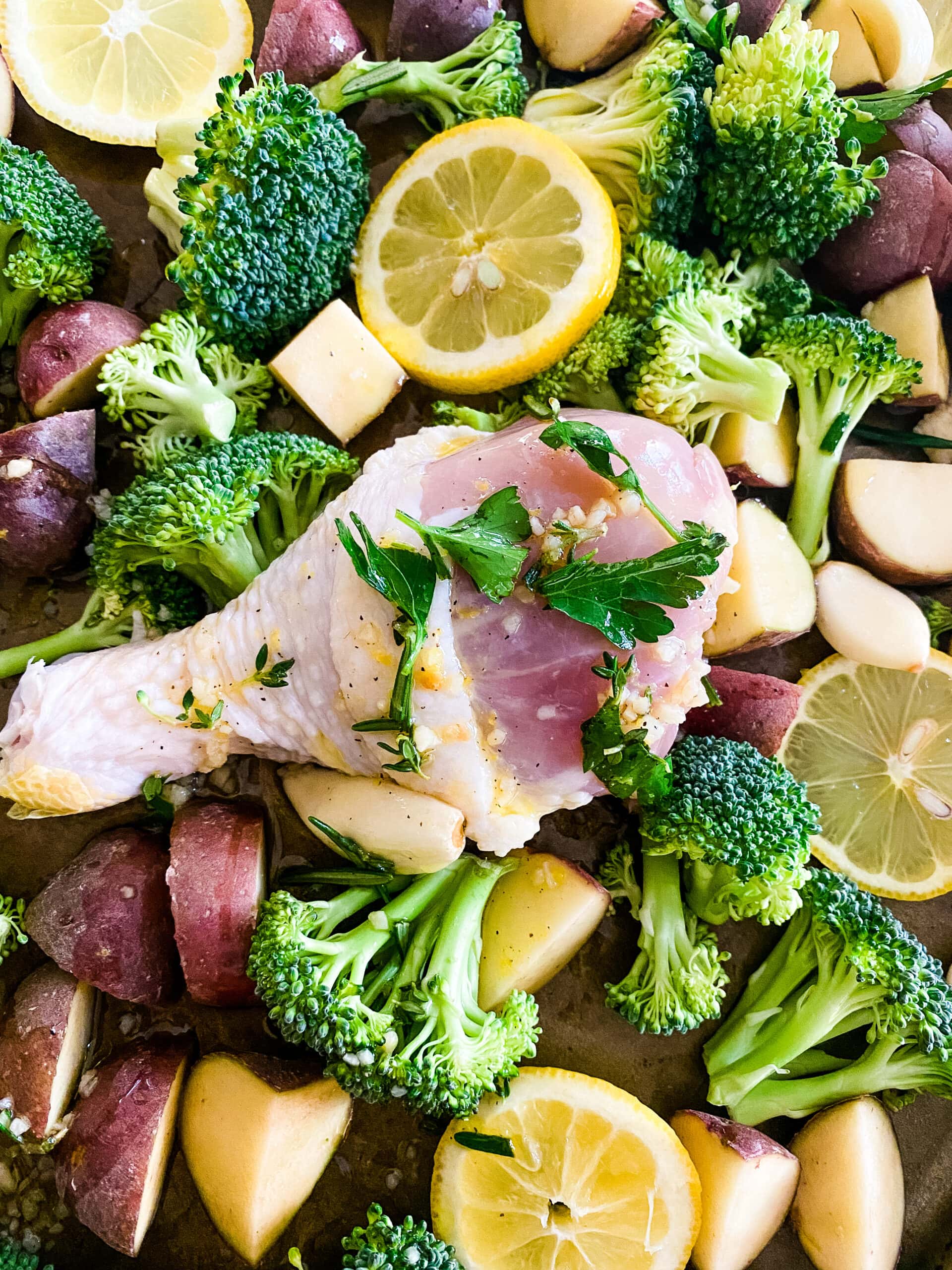 A closeup view of a sheet pan with chicken drumsticks, broccoli florets, baby red potatoes, lemon slices, and garlic cloves for Sheet Pan Lemon Garlic Chicken. Cooking With Fudge