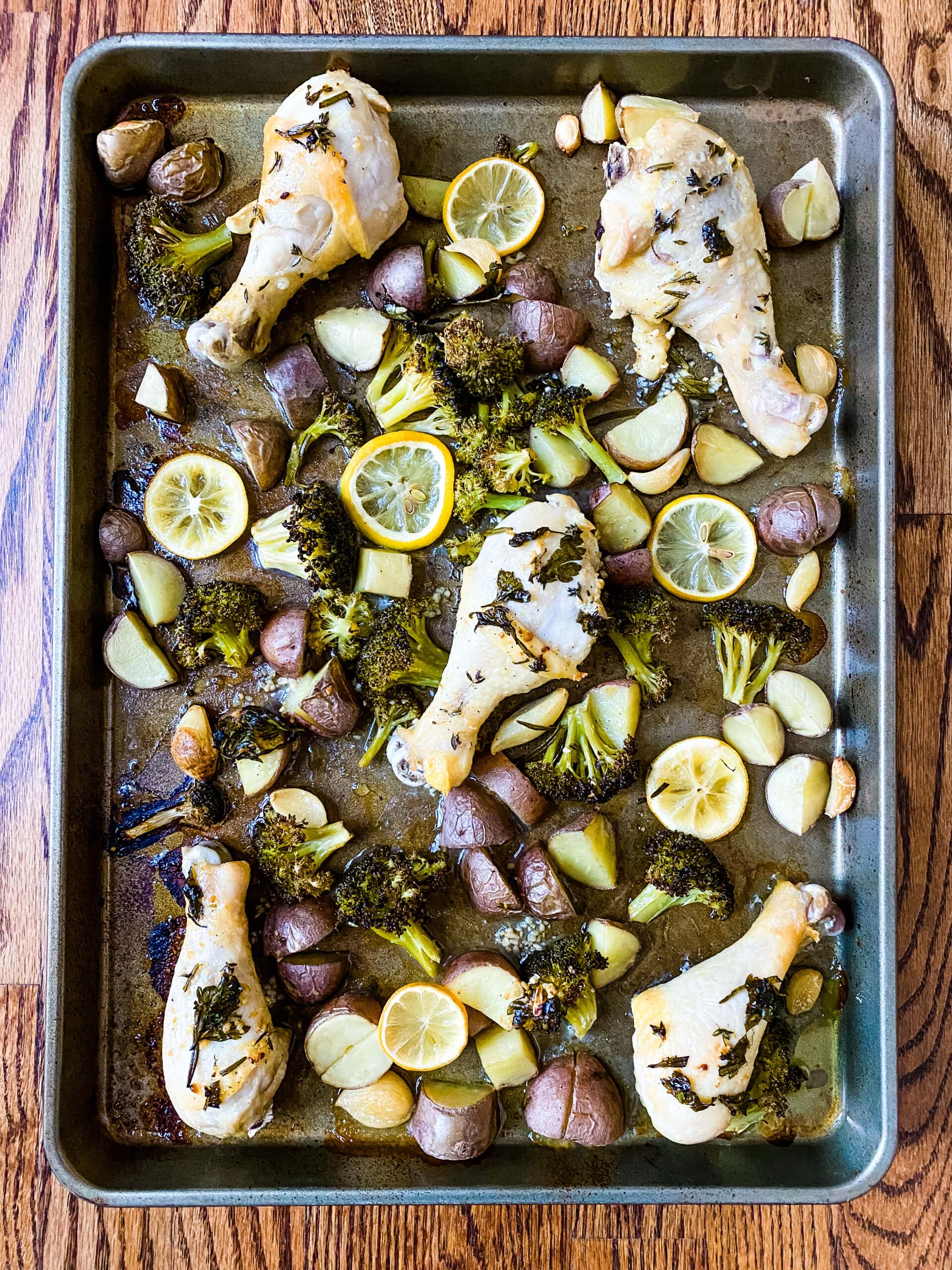 A sheet pan with chicken drumsticks, broccoli florets, baby red potatoes, lemon slices, and garlic cloves for Sheet Pan Lemon Garlic Chicken. Cooking With Fudge