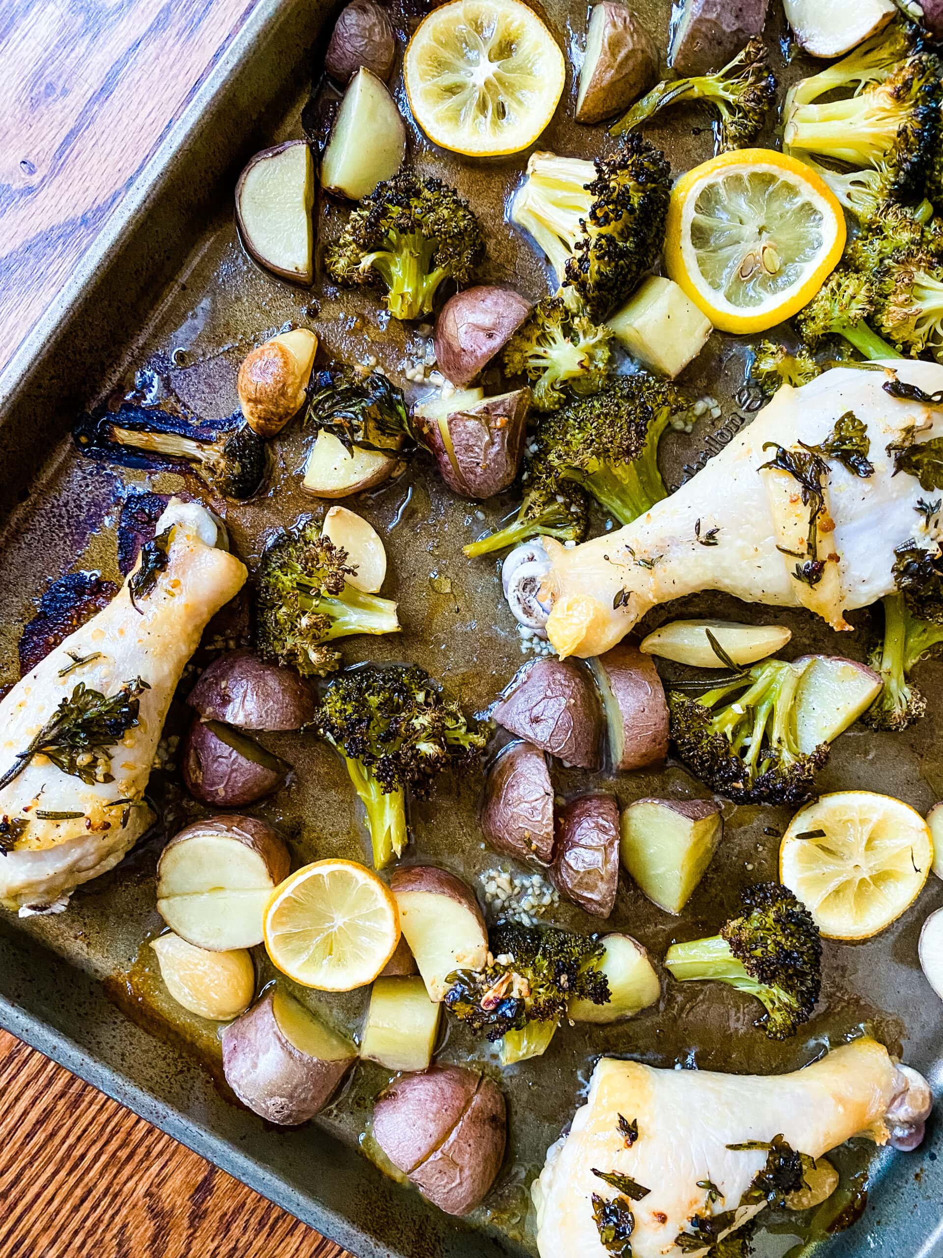 A sheet pan with chicken drumsticks, broccoli florets, baby red potatoes, lemon slices, and garlic cloves for Sheet Pan Lemon Garlic Chicken. Cooking With Fudge